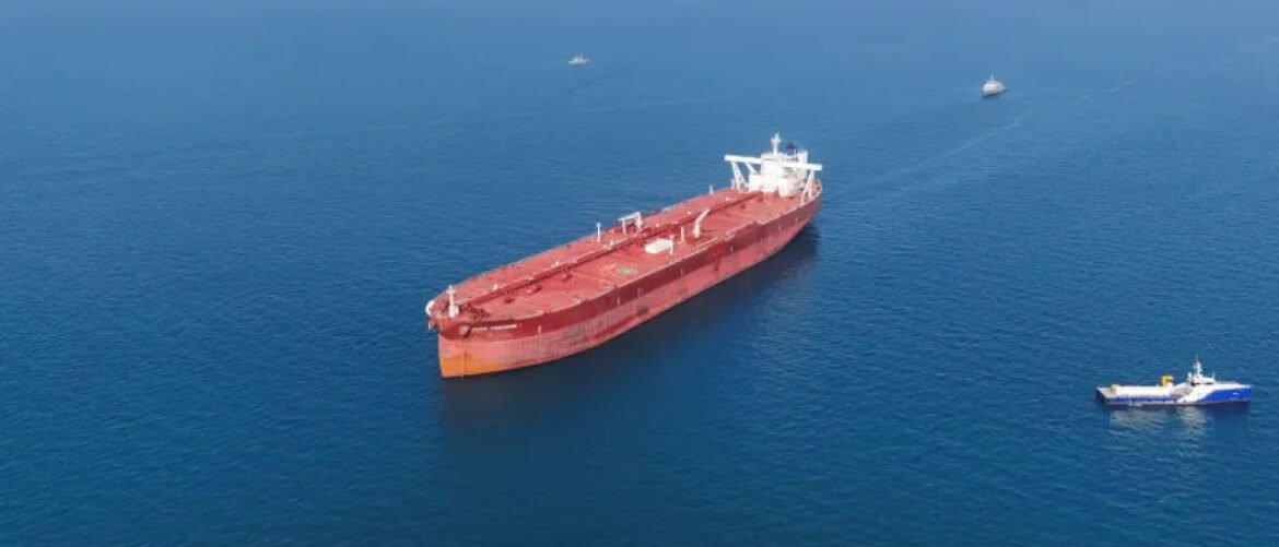 Aerial view of a large red oil tanker sailing in calm blue water with two small support boats nearby.