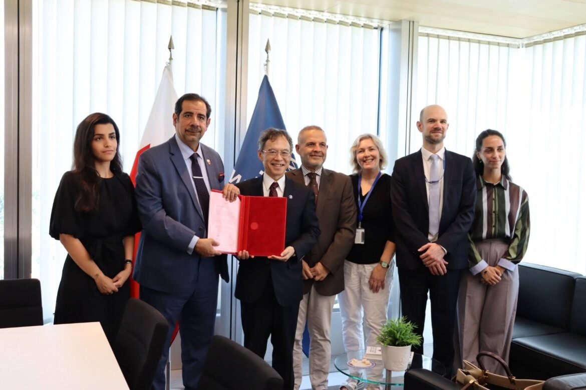 Six professionals in business attire posing in a conference room; one man in a blue suit holds a red folder while the others smile for the photo, with flags in the background.