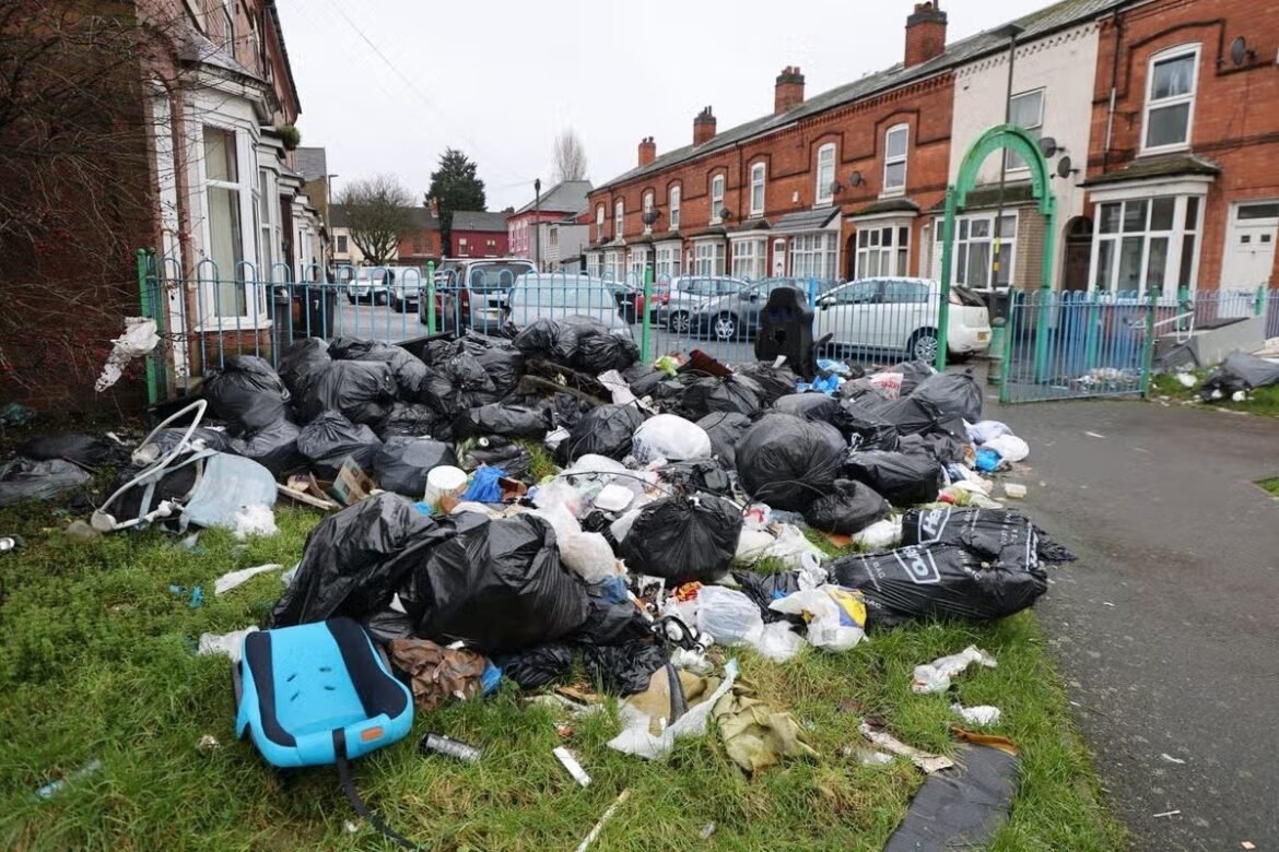 Large pile of black garbage bags and scattered litter on a grassy verge beside a residential street with brick houses and parked cars behind a blue fence.