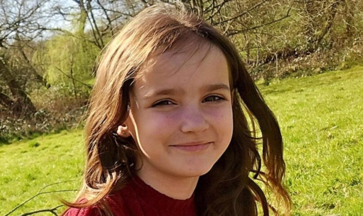 Smiling young girl outdoors in a sunny park with green grass and trees, wearing a red shirt.