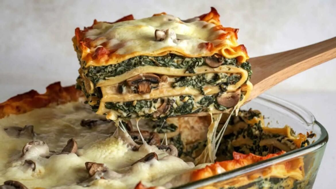 Lasagna layered with spinach, mushrooms and melted cheese being lifted from a baking dish with a wooden spoon edge visible.