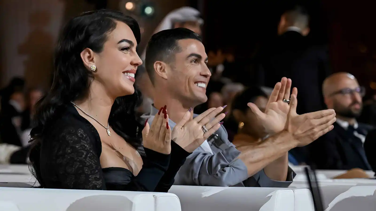 Smiling Woman in a Black Lace Top and Necklace Claps Beside a Man Both in an Audience at an Event with Other Attendees in the Background  Aleumumnews