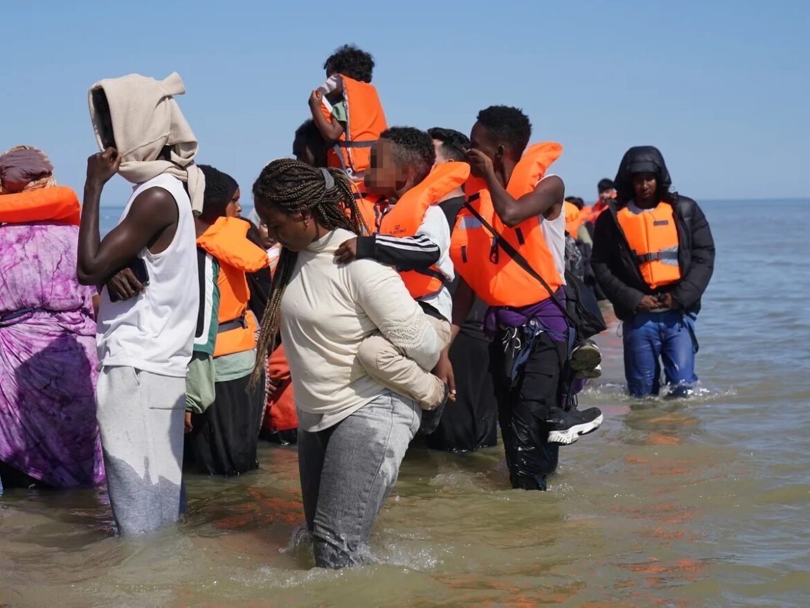 Group of people wearing orange life jackets wading through shallow coastal water, some helping to carry a person forward.