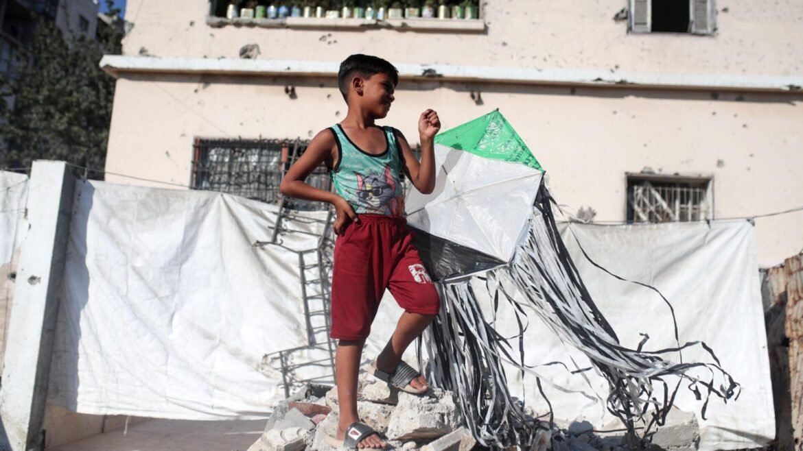 Boy stands on rubble, holding a green and white kite with long black streamers.