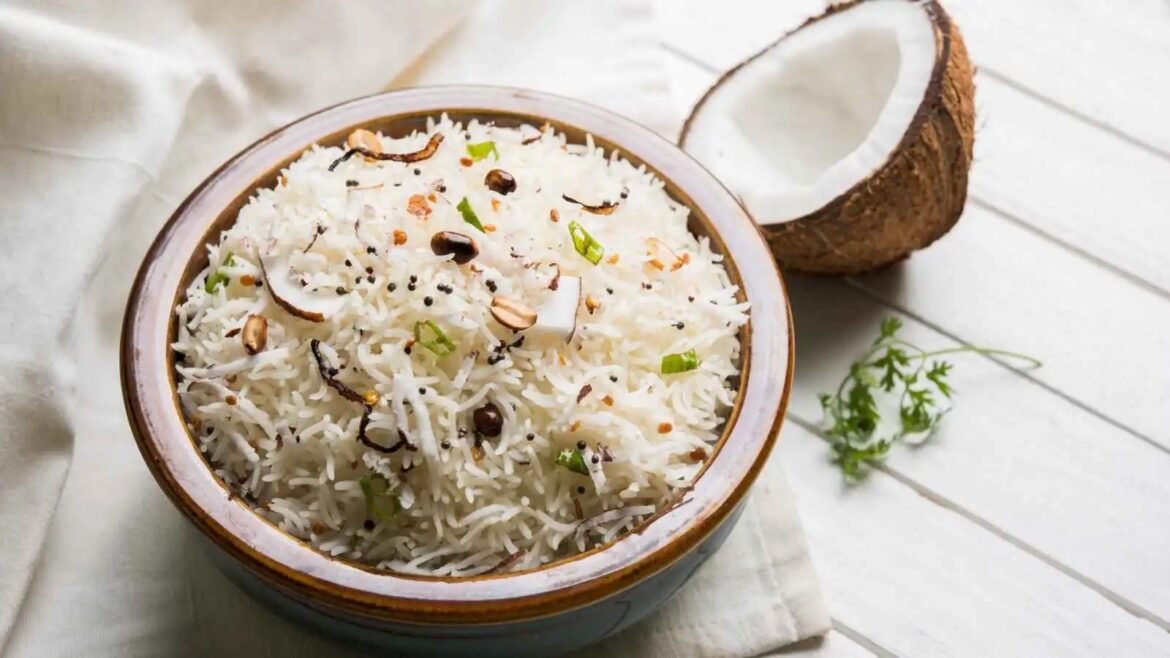 Bowl of fluffy coconut rice with fried coconut, green onions, and sesame seeds, accompanied by a halved coconut on a white wooden surface.
