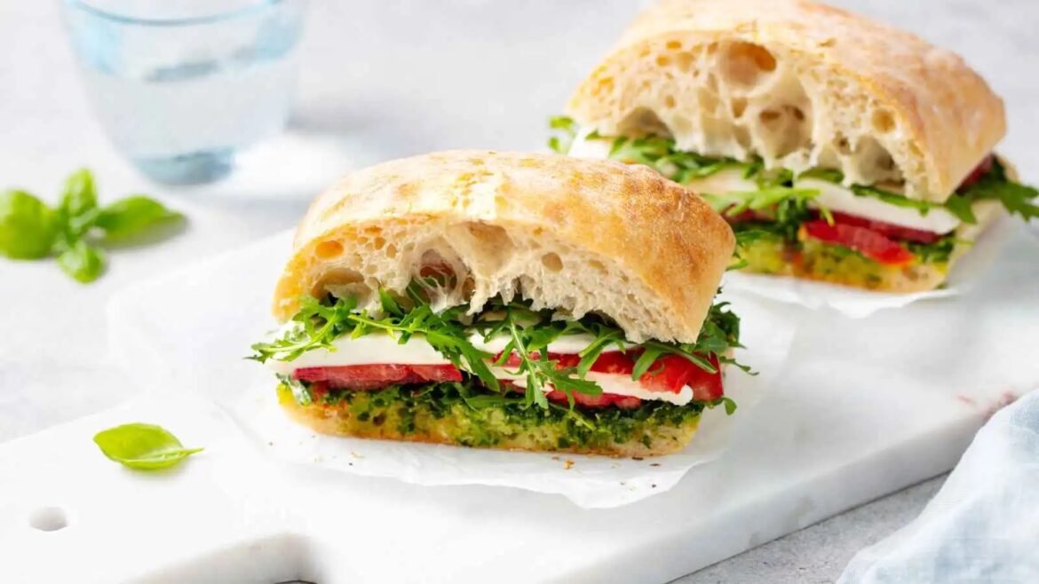 Two halves of a rustic ciabatta sandwich filled with mozzarella, tomato, arugula, and green pesto on a white surface.