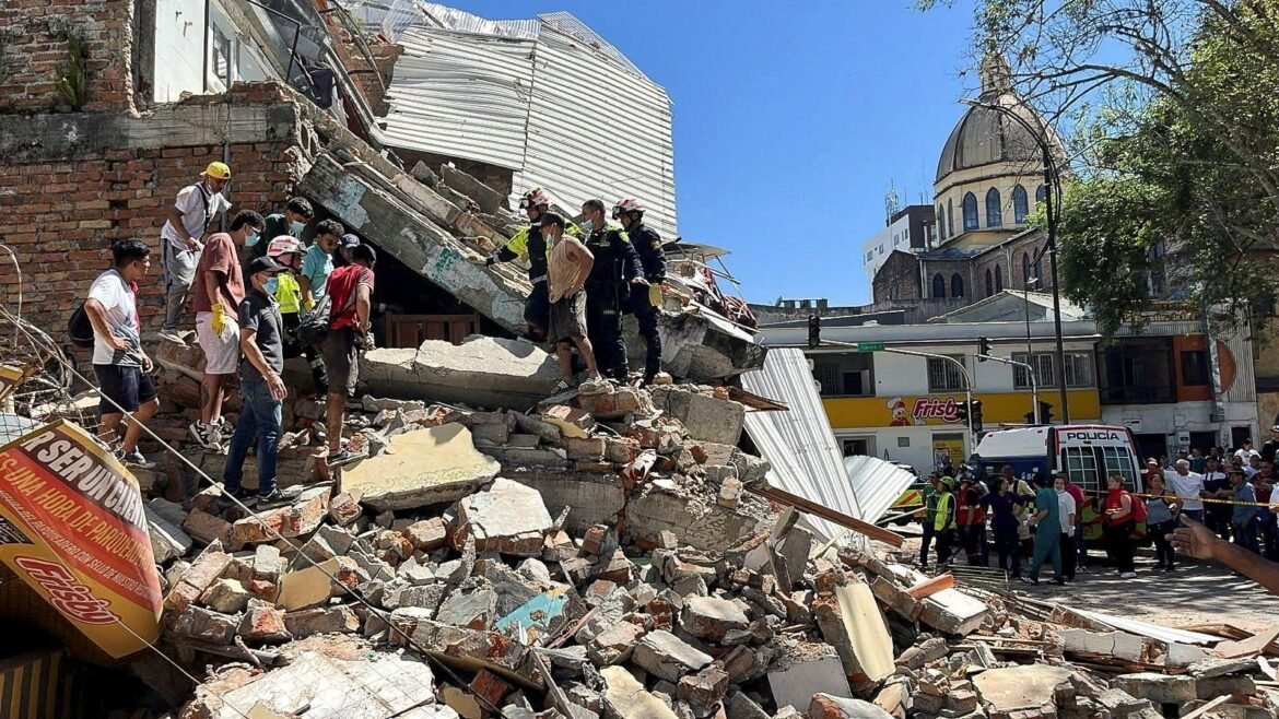 Rescuers in helmets and uniforms stand on rubble of collapsed building, surveying debris as bystanders watch nearby