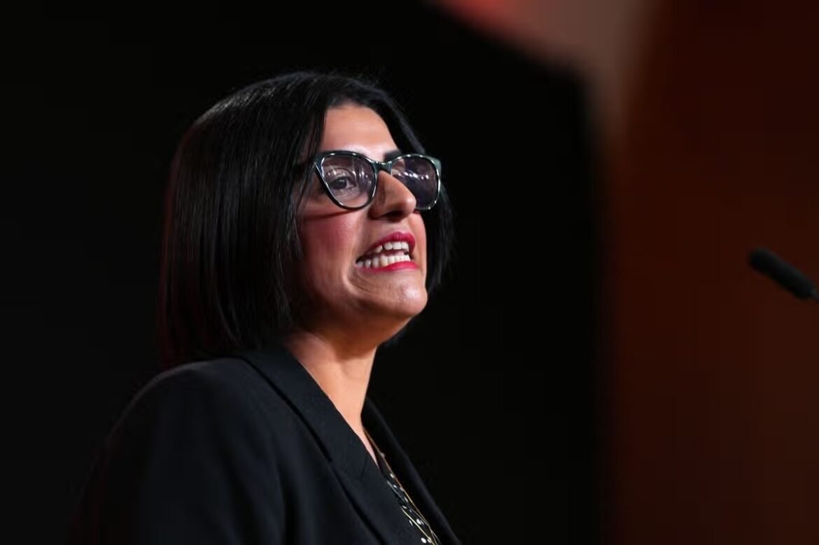 Female speaker wearing glasses and a black blazer delivering a talk on stage against a dark background.