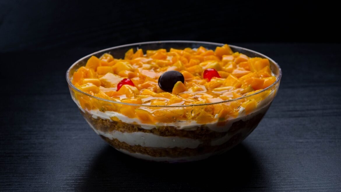 Layered mango dessert in a glass bowl with mango chunks, cream, and crumb crust, topped with a cherry and yellow fruit pieces.