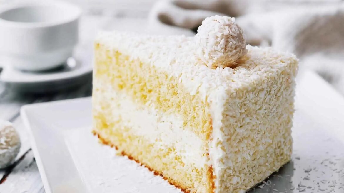Slice of yellow coconut cake with white frosting and shredded coconut on the outside, topped with a small curled coconut piece on a white plate.