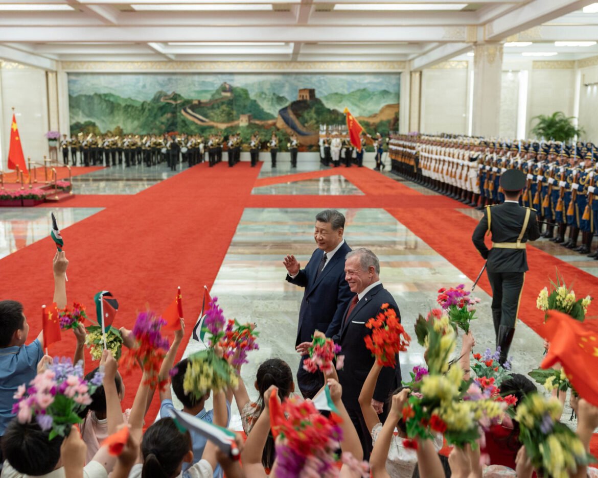 Crowded ceremonial hall with soldiers in uniform lining the red carpet and people waving colorful flowers as officials stand at the center behind a flag.