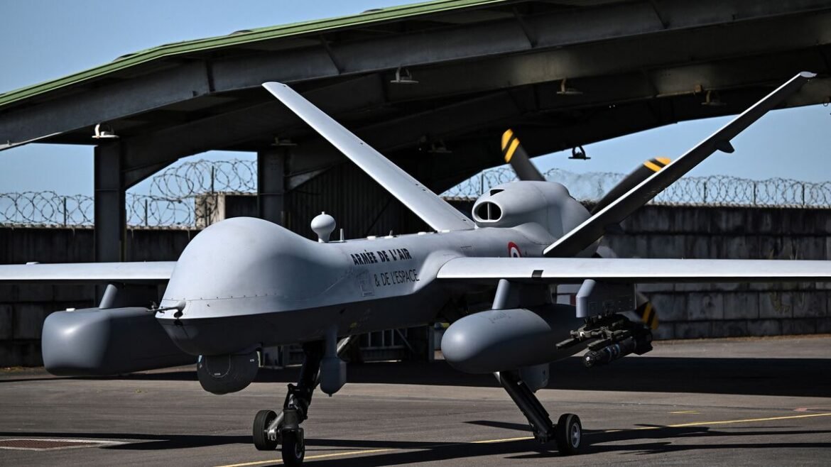 French Air Force unmanned drone parked on the tarmac beside a hangar, with barbed fencing in the background, conveying its military use and location.