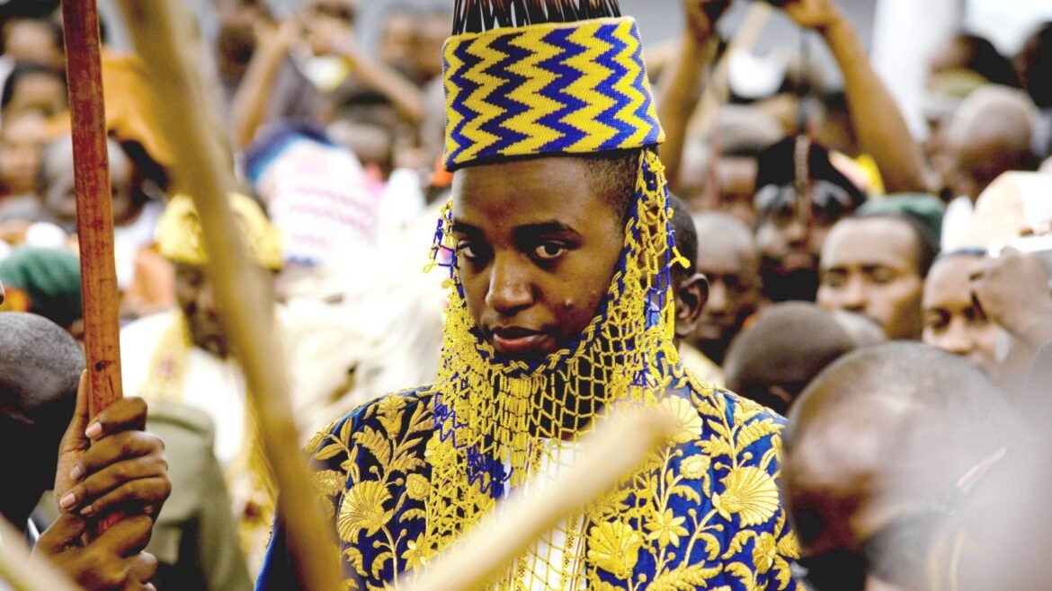 Young man in blue and gold traditional ceremonial attire with a tall patterned cap, standing in a crowd at a festival.