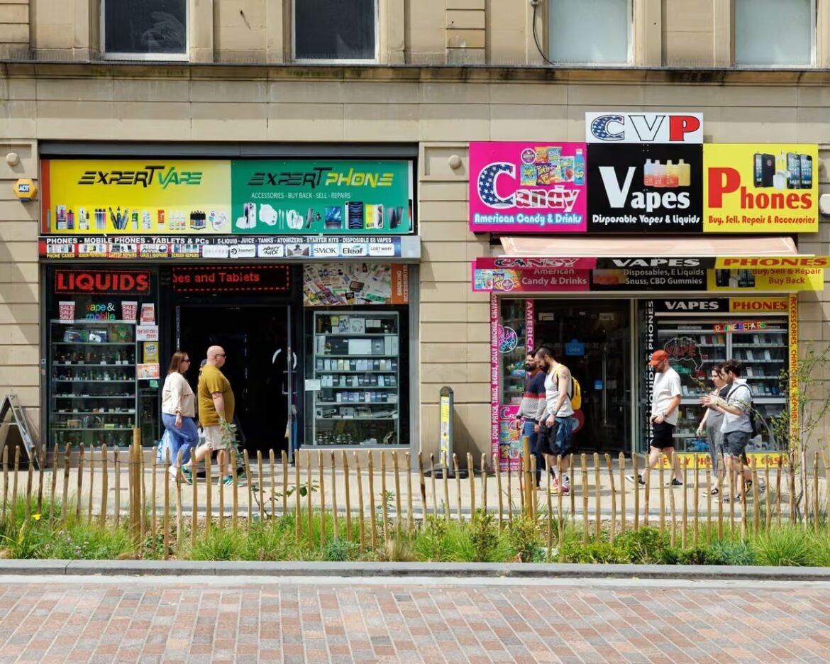 Pedestrians walk past a row of colorful vape, phone, and candy shop storefronts on an urban street.