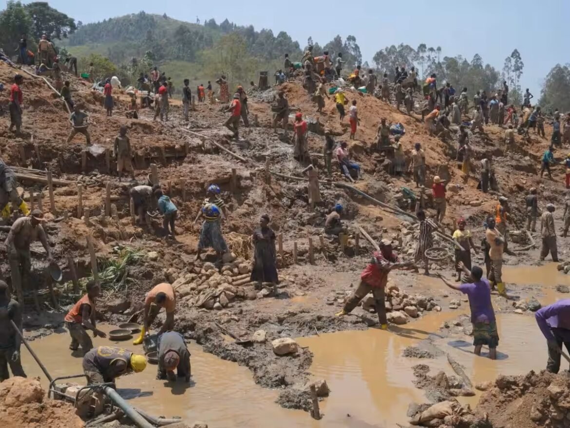 Large group of people digging and sorting through a muddy open-pit site on a hillside, using buckets and makeshift tools near muddy water.