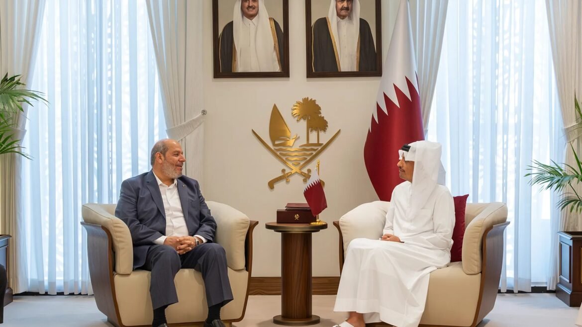 Two men sit and talk in a formal room; a large Qatar flag and gold emblem are displayed behind them with portrait photos above.