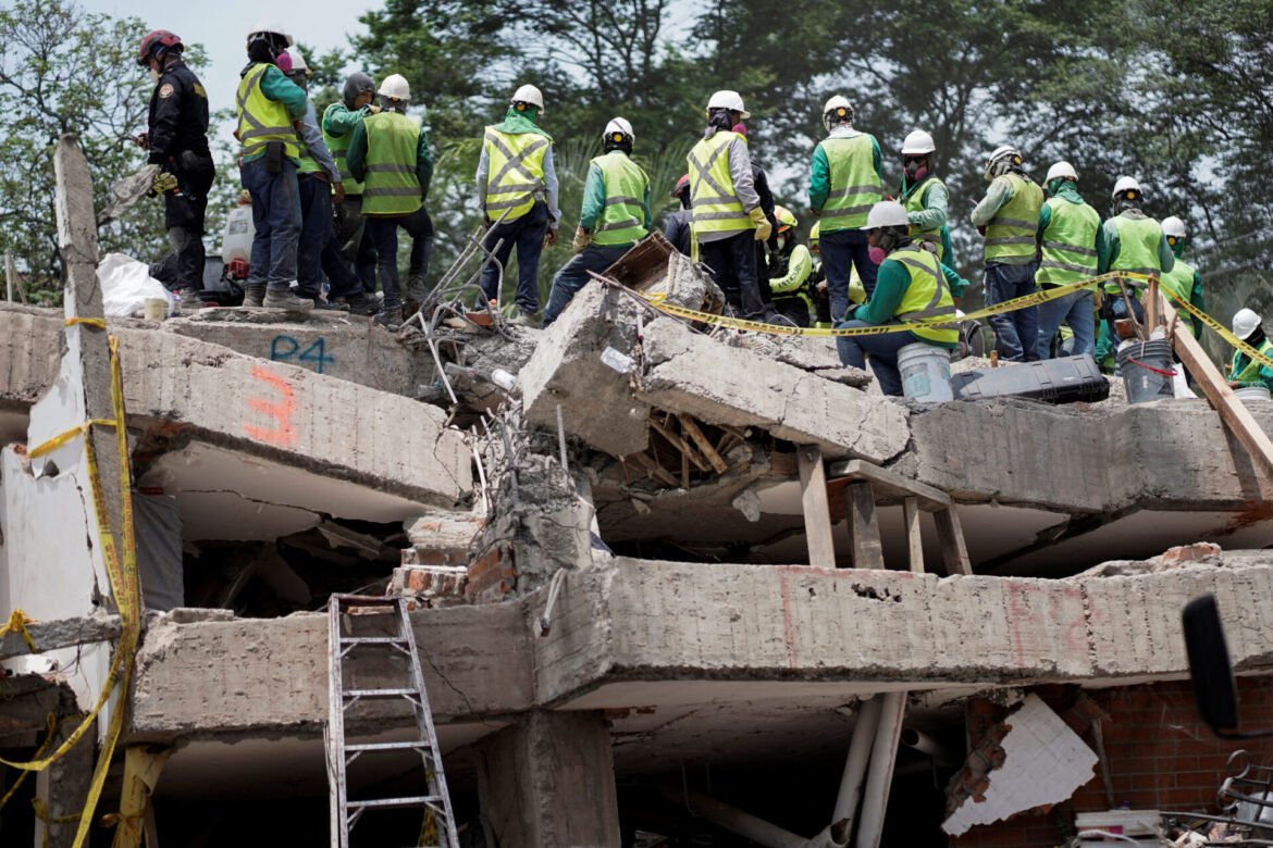 Group of construction workers in safety vests and hard hats on a collapsed concrete structure with yellow caution tape in the foreground or around the site: a rubble-filled work zone.
