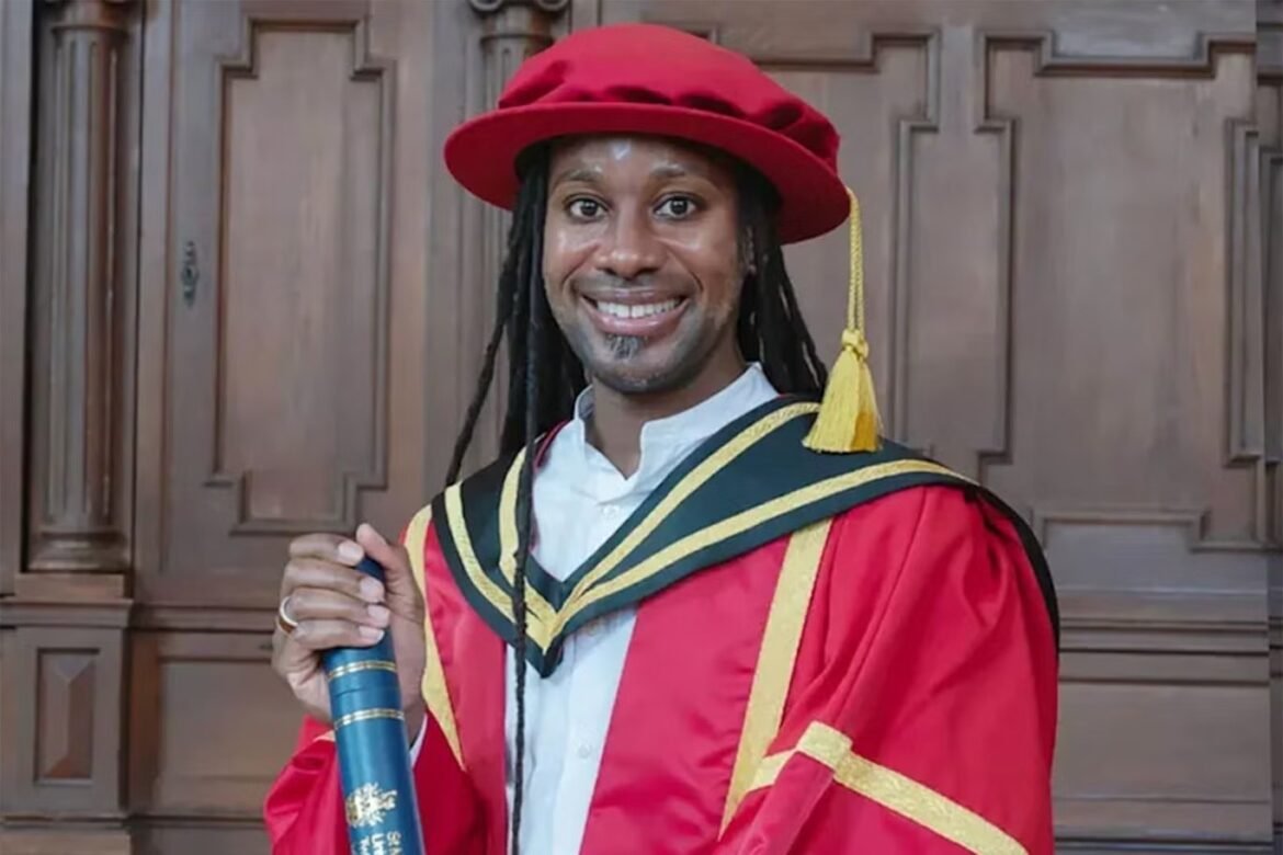 Graduate in a red cap and gown holding a rolled diploma, smiling at the camera in front of wooden panels.