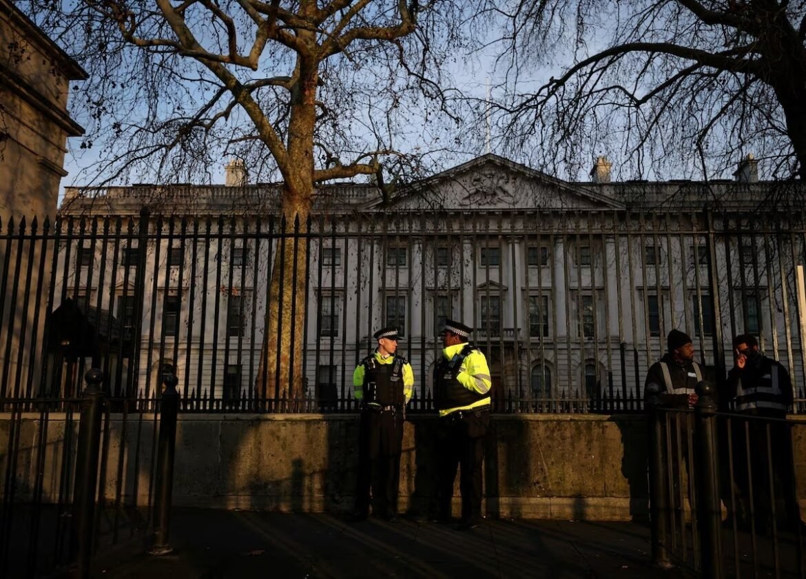 Two police officers in neon jackets stand and chat beside a tall iron fence in front of a large, historic building with leafless trees overhead.