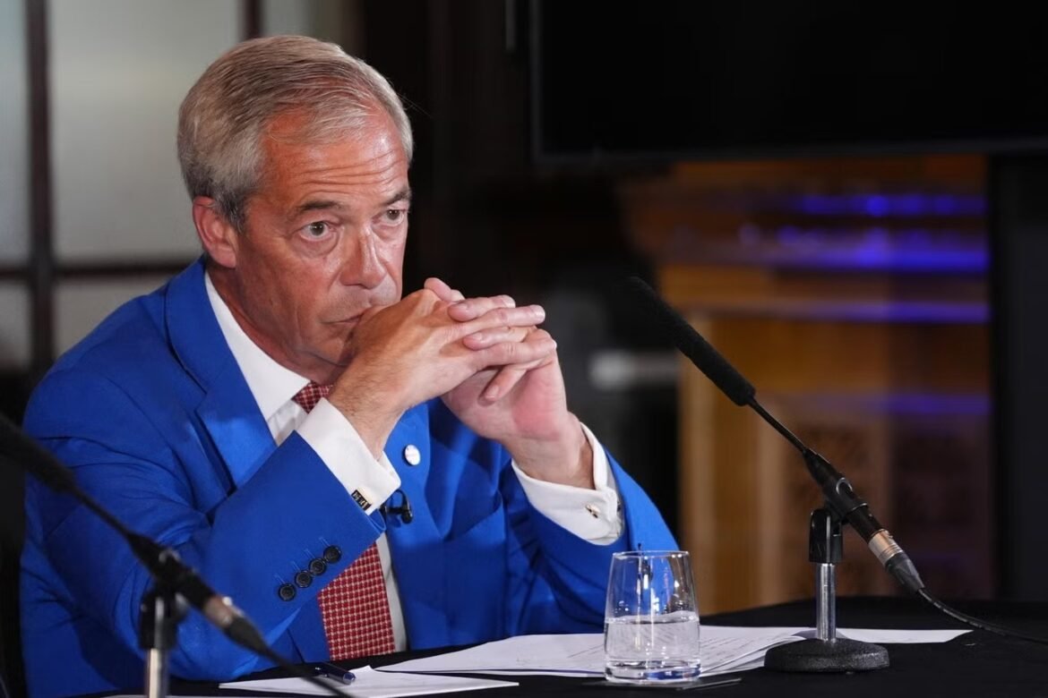 Man in a blue suit sits at a panel, hands clasped under his chin, microphones in front, a glass of water on the table.