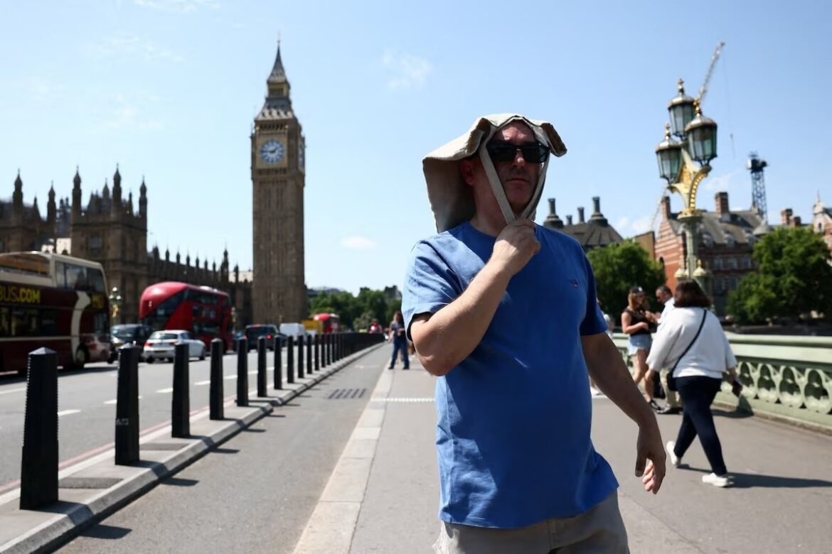 Man in a blue T-shirt with a cloth over his head and sunglasses walks on Westminster Bridge, with Big Ben and Parliament in the background.