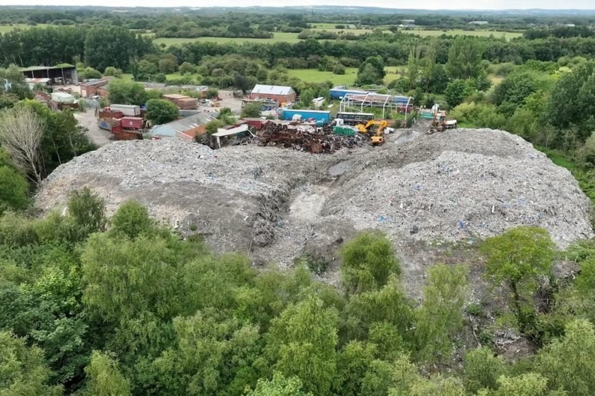 Aerial view of a large rubble mound and construction debris with excavators at a scrapyard, bordered by trees and fields.