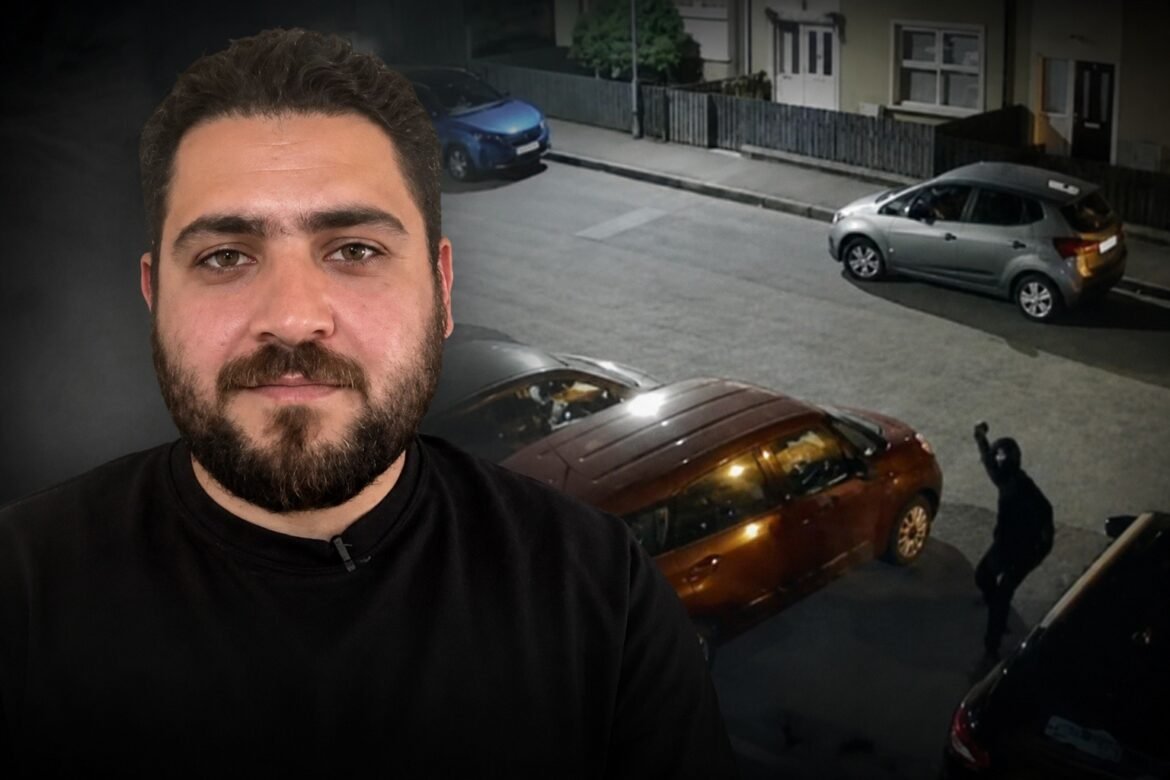 Close-up portrait of a bearded man in a black shirt; a street with parked cars and a car crash is visible behind him on the right side of the image.