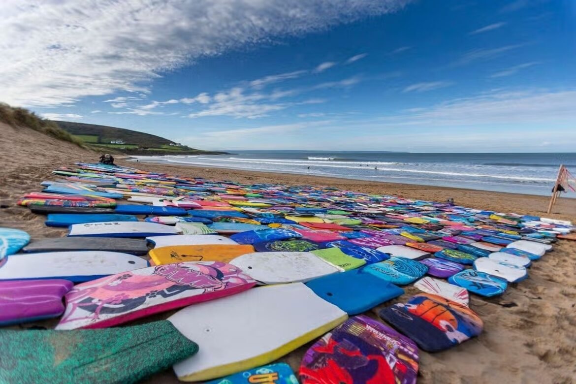 Colorful surfboards and bodyboards laid out on a sandy beach with the ocean and blue sky in the background.