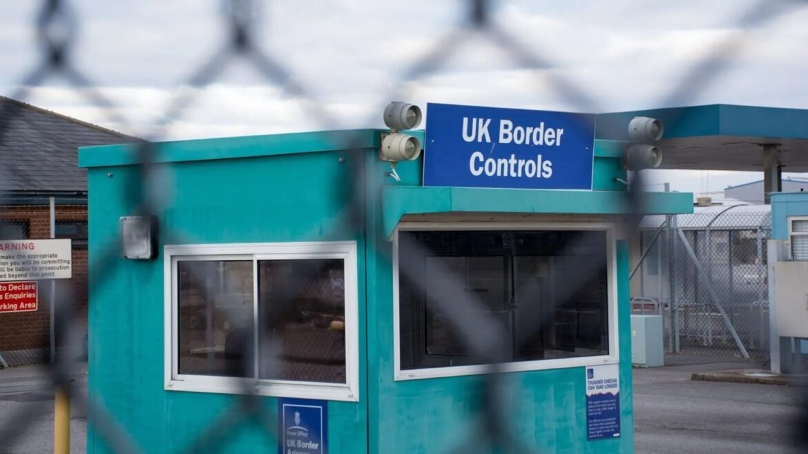 Teal border control booth with a blue 'UK Border Controls' sign, viewed through a chain-link fence.