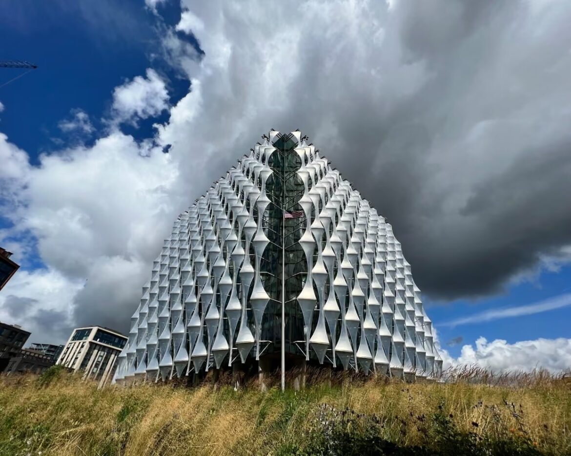 Pyramid-shaped glass-and-metal building with a honeycomb-like white facade, rising from a grassy field under a dramatic cloudy sky.
