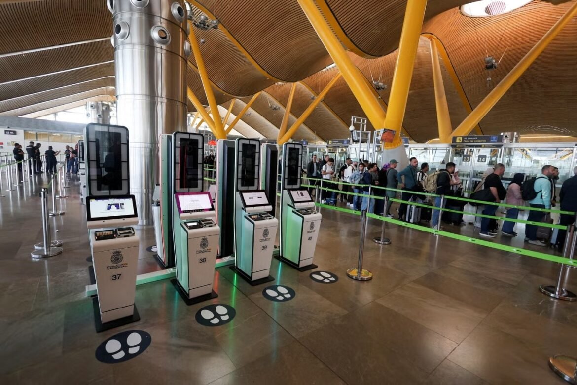 Modern airport terminal with a row of self-service check-in kiosks and passengers waiting in line behind green ropes, yellow structural beams overhead.