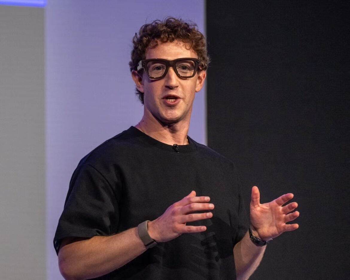 Male speaker with curly hair and dark glasses delivering a talk on stage, gesturing with hands.