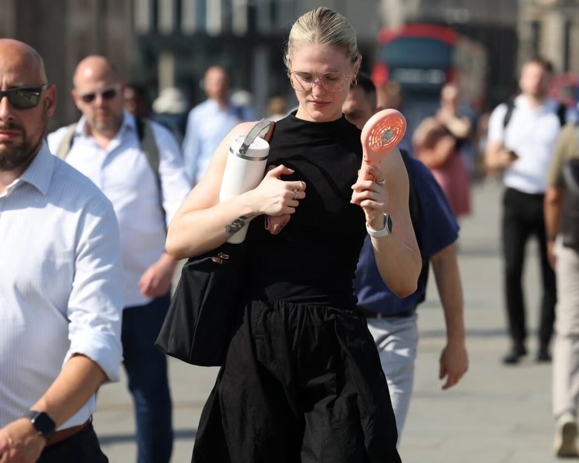 Woman in a black sleeveless dress walks through a crowded urban street, carrying a white water bottle and a pink handheld fan as others pass by.