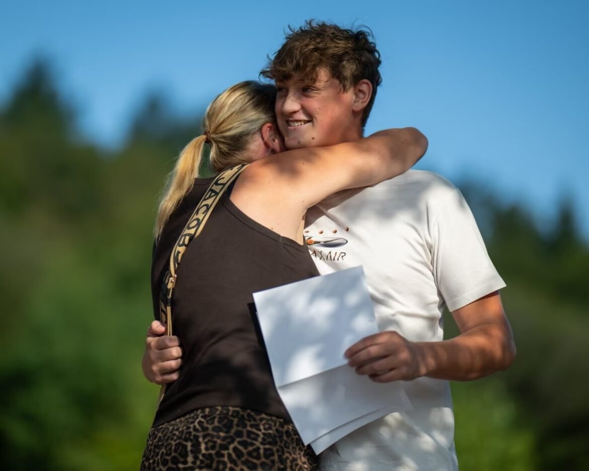 Two people share a hug outdoors, a smiling man in a white t-shirt holds a stack of papers while a woman in a black top and leopard-print skirt embraces him.