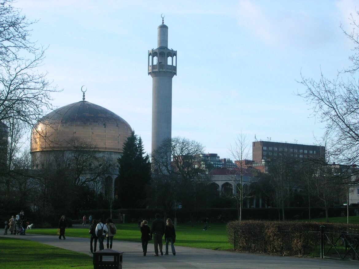 Park scene with a gold-domed mosque and tall minaret in the background; people walk along a path among bare trees.