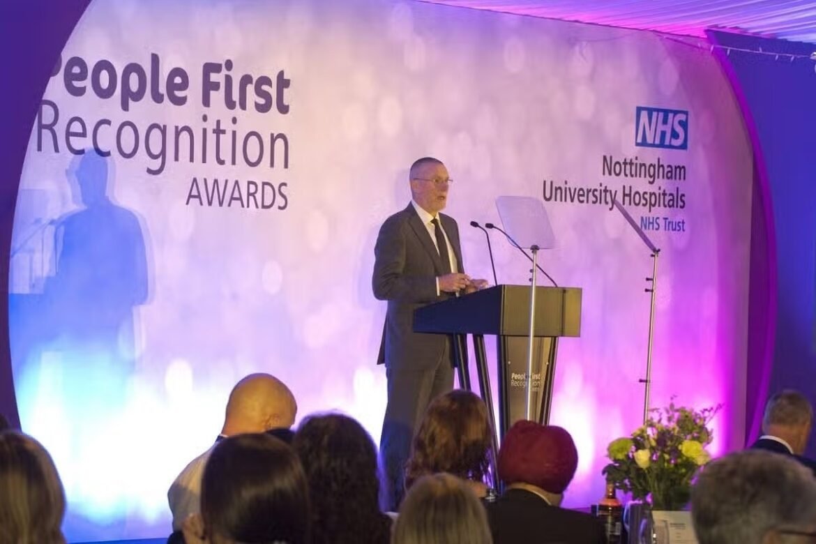 Speaker at a podium delivering a address at the People First Recognition Awards stage, NHS signage visible in background with purple lighting