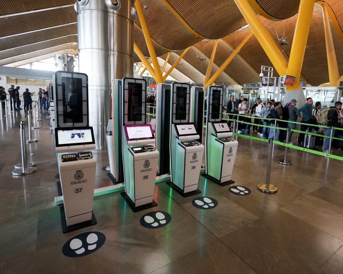 Row of self-service airport check-in kiosks with large screens and numbered stands, passengers queuing in the background near yellow structural beams.