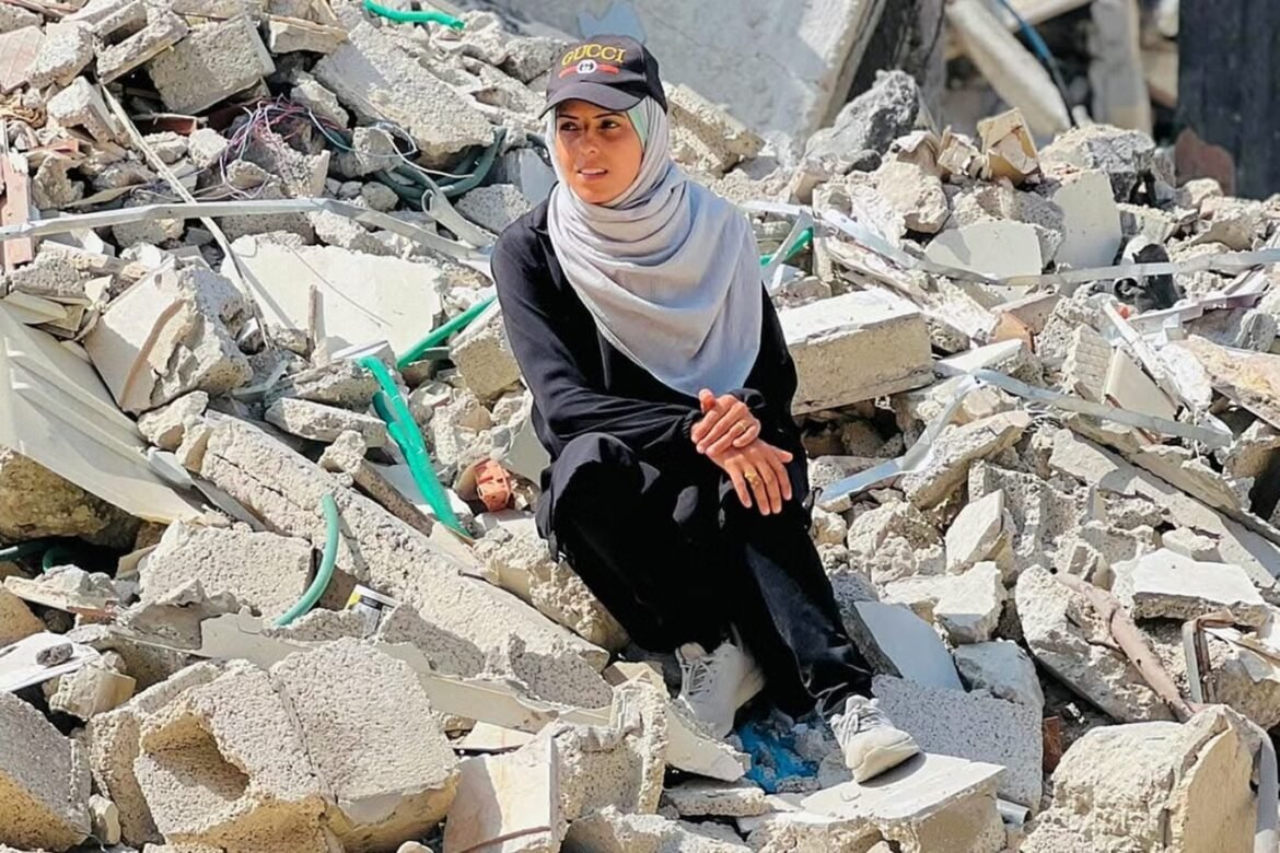 Woman wearing a gray hijab and black clothing sits on a mound of rubble from a collapsed building, looking thoughtful.