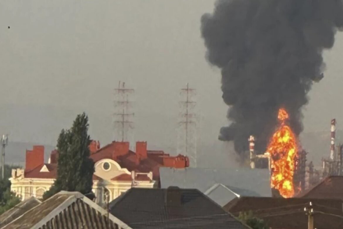 Industrial facility on fire with large orange flames and thick black smoke rising, seen behind a row of houses in a residential neighborhood.
