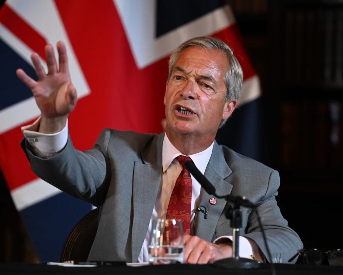 Man in a gray suit and red tie speaks into a microphone while gesturing with his raised left hand, with a British flag in the background.