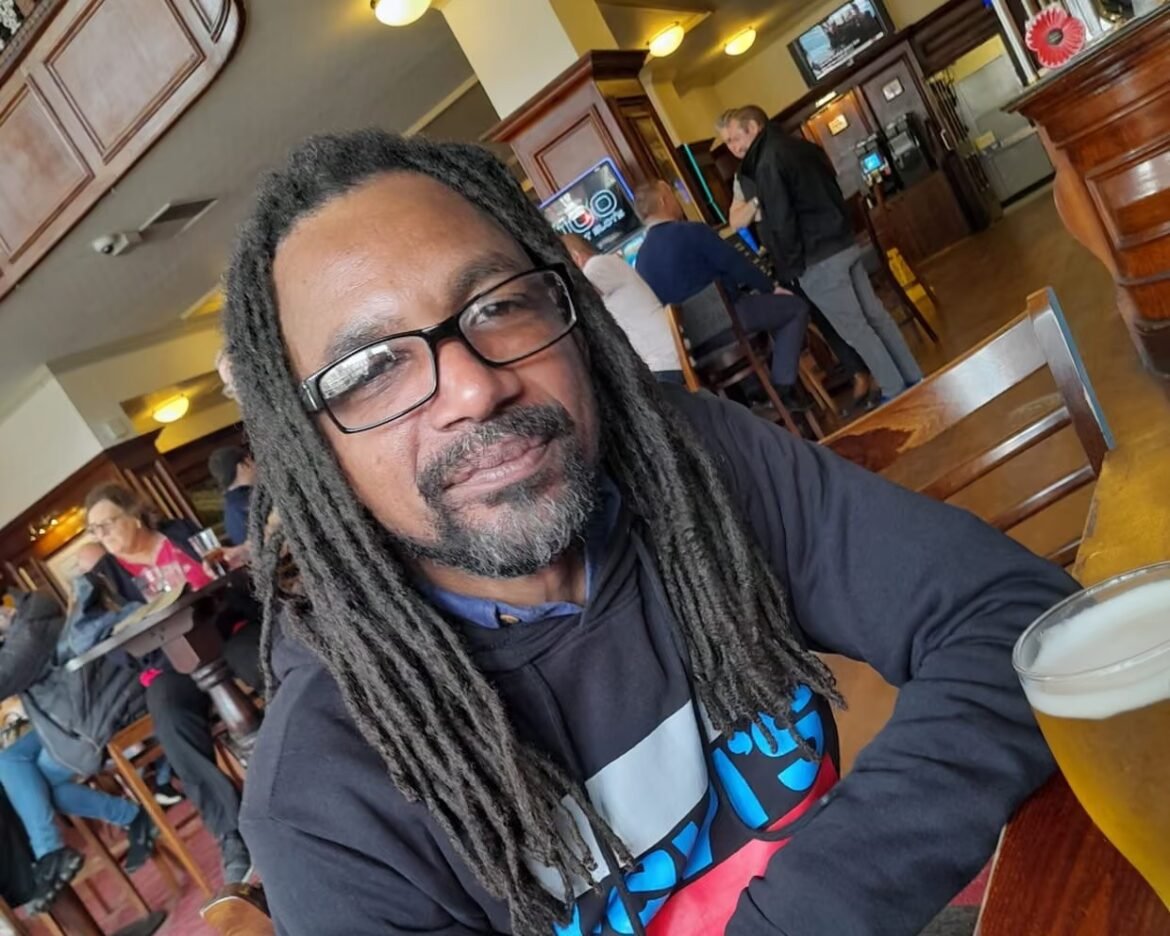 Man with glasses and long dreadlocks sits at a wooden table in a pub, smiling, with a pint of beer in the foreground and other patrons in the background.