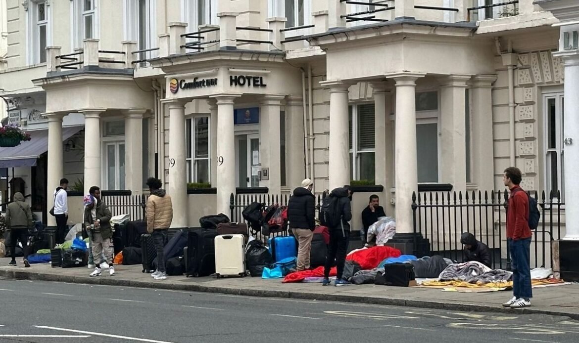 People with suitcases and blankets lined up along the sidewalk outside a hotel, suggesting a temporary encampment or travel scene.