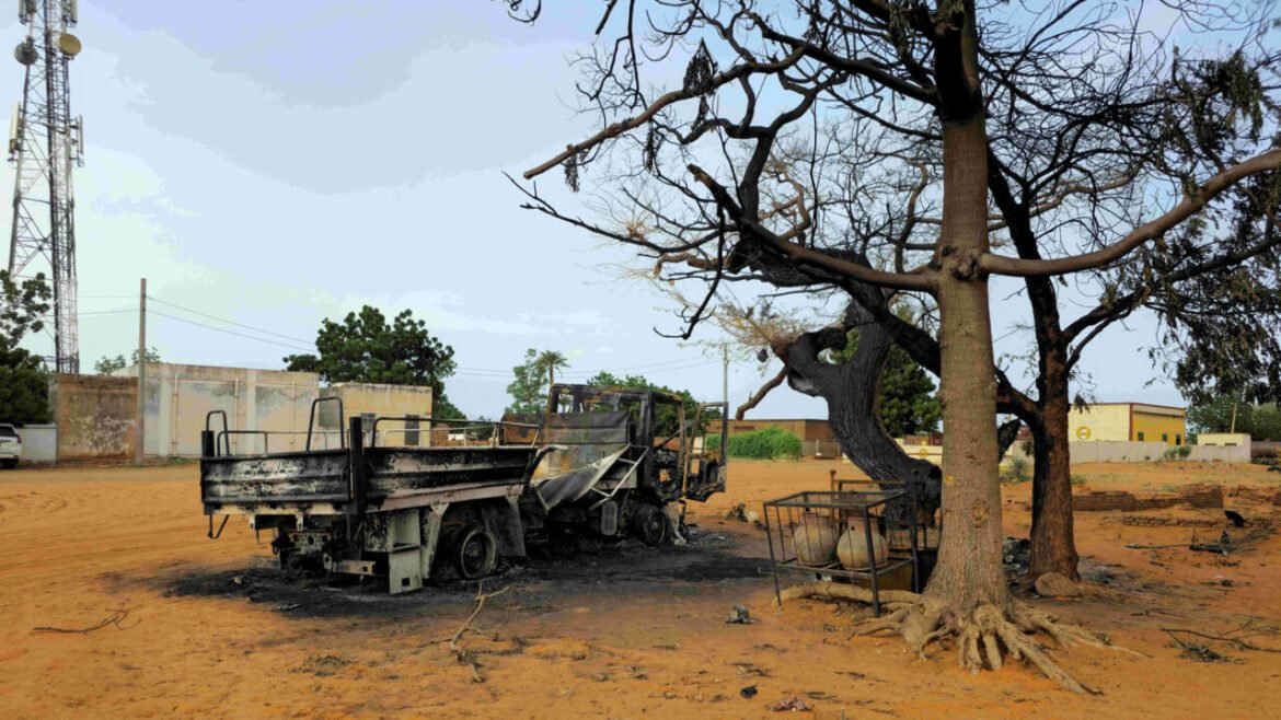 Charred dump truck in a sandy lot next to a burnt, leafless tree; debris scattered around.