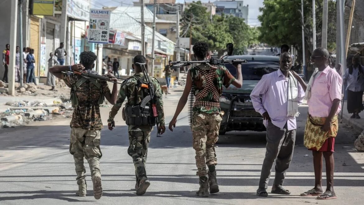 Four armed soldiers walk down a street, rifles resting on their shoulders while civilians watch nearby.