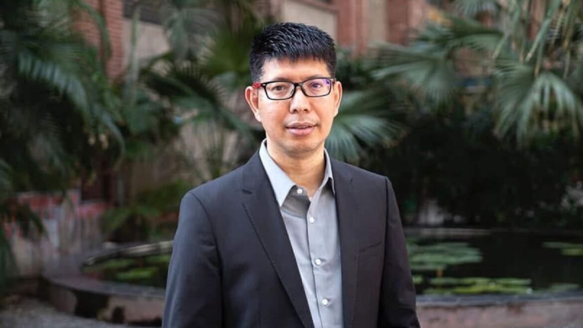 Professional man in glasses wearing a dark suit and light gray shirt, posing outdoors with greenery and a pond in the background.