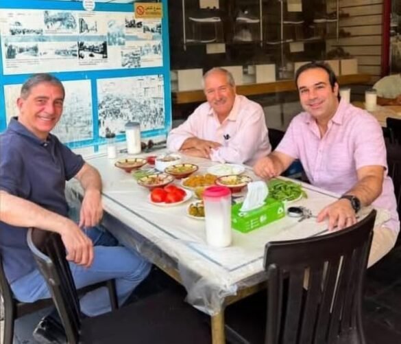 Three men sit at an outdoor cafe table sharing a meal, bowls of food and tomatoes in front, all smiling at the camera.