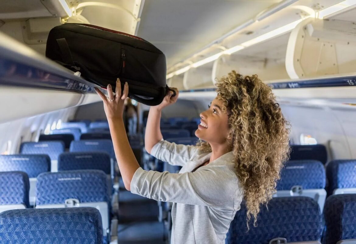 Passenger places a black backpack into the airplane overhead bin as the cabin seats are visible in the background.
