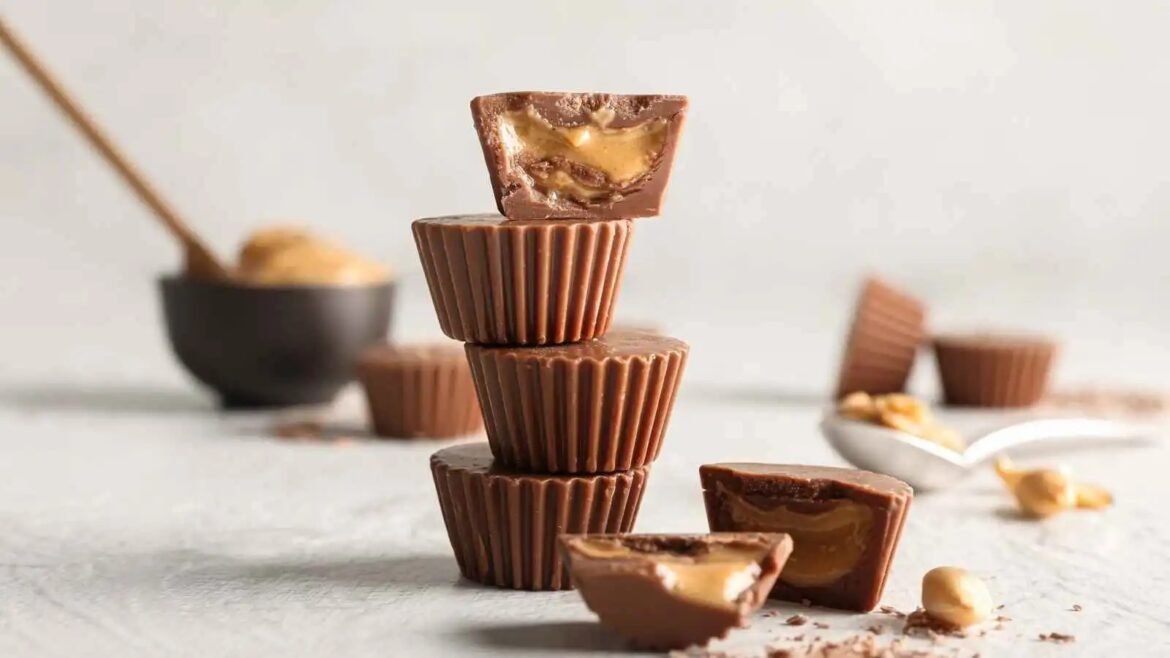 Stack of four chocolate peanut butter cups on a light surface, with a broken piece showing the creamy filling and scattered peanuts nearby.