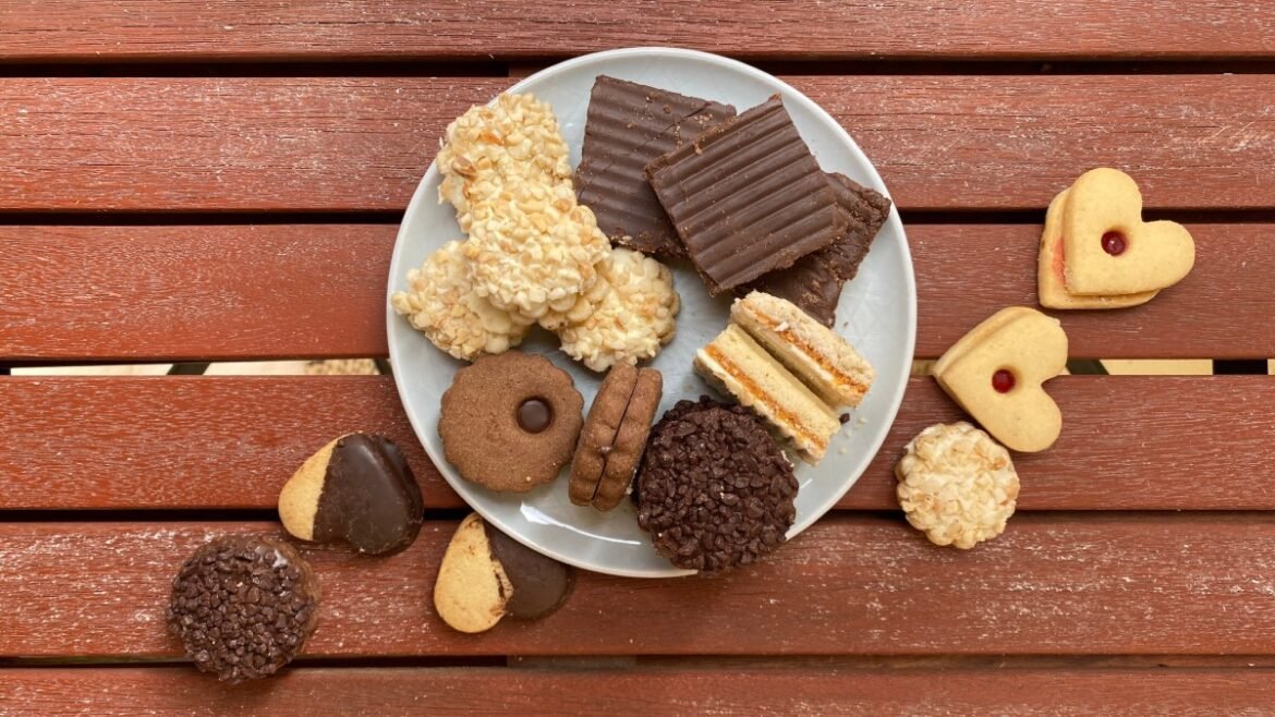 Assorted cookies on a white plate atop a red wooden table, including chocolate bars, almond cookies, sandwich cookies, and heart-shaped cookies with jam centers.
