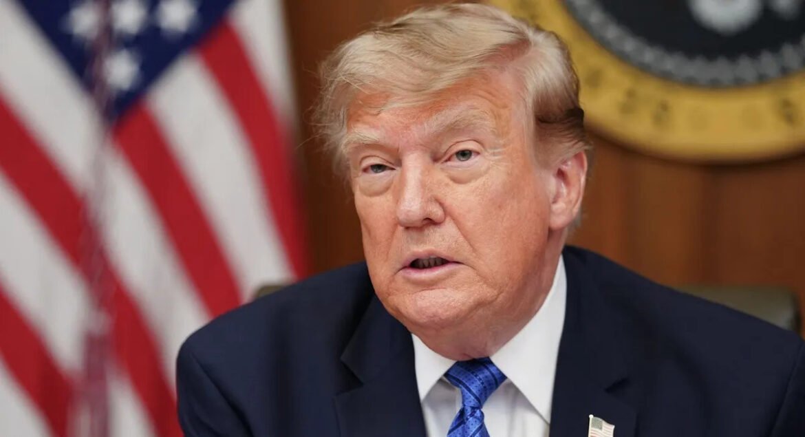Close-up of a man in a navy suit and blue tie, with an American flag in the background.